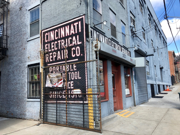 Außenansicht des Cincinnati Electrical Repair Co.-Gebäudes mit einer Schautafel, einem Metallzaun, einem Müllbehälter, Strommasten, Drähten, Bäumen und einem bewölkten Himmel.