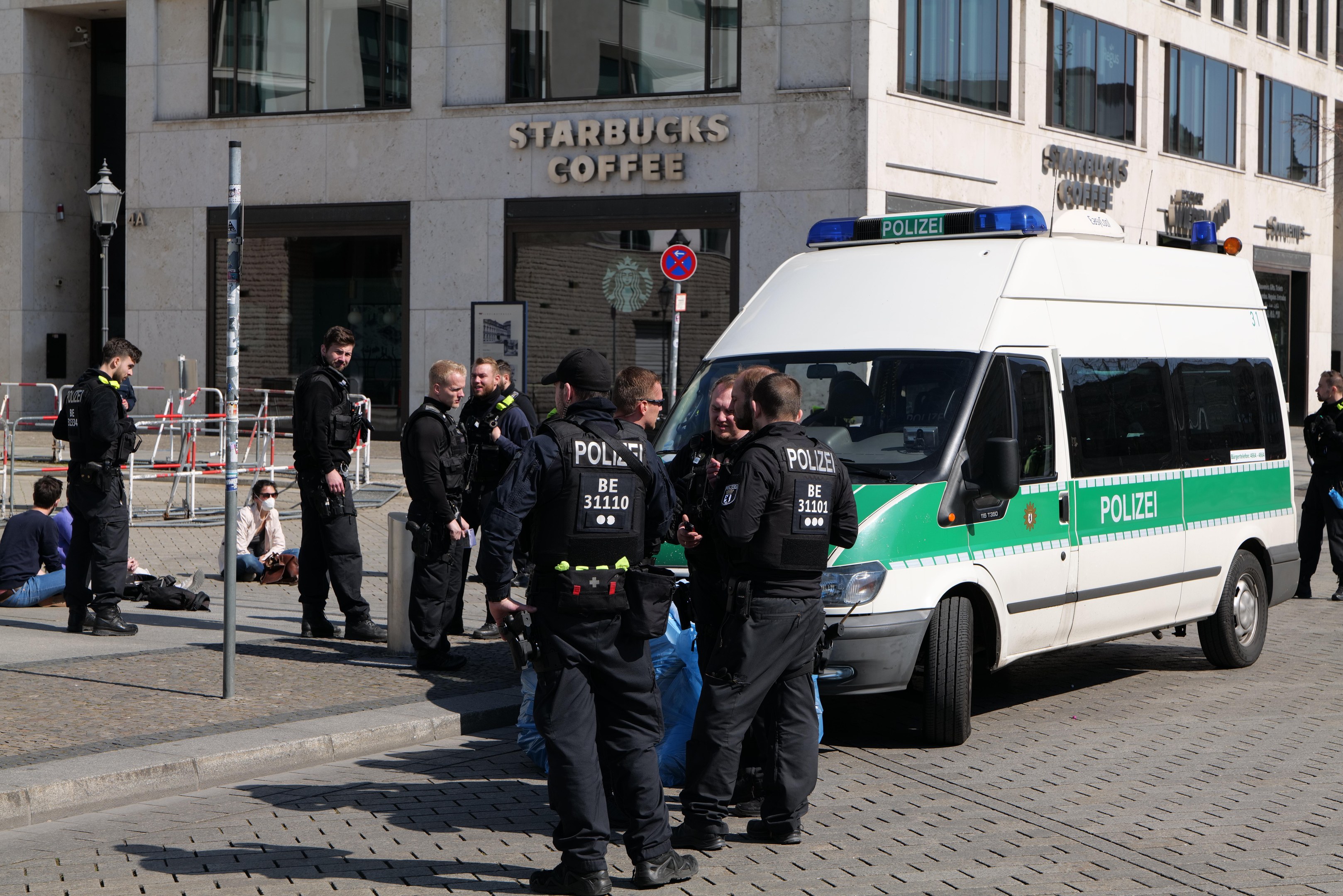 Eine Gruppe von Polizisten steht vor einem Starbucks-Café, rechts daneben ein Van und links ein paar Menschen; im Hintergrund ist ein Gebäude mit Fenstern, ein Schild, ein Laternenmast und ein Zaun zu sehen.