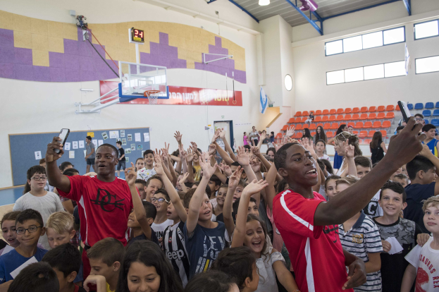 Gruppe von Kindern vor einem Basketballfeld mit Mobiltelefonen, einer Tafel, einer Uhr, einem Torpfosten, einem Basketballkorb, Deckenleuchten, Stühlen und Fenstern im Hintergrund.