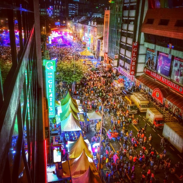 Eine volle Karnevalsstimmung mit Zelten, Geschäften, Gebäuden und Bannern, mit beleuchteter Fläche im Hintergrund.