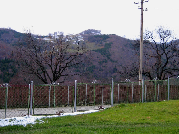 Eine grüne Landschaft mit einem Zaun, einem Pfahl und Bäumen, mit ein paar Häusern im Hintergrund auf einem Hügel sichtbar.