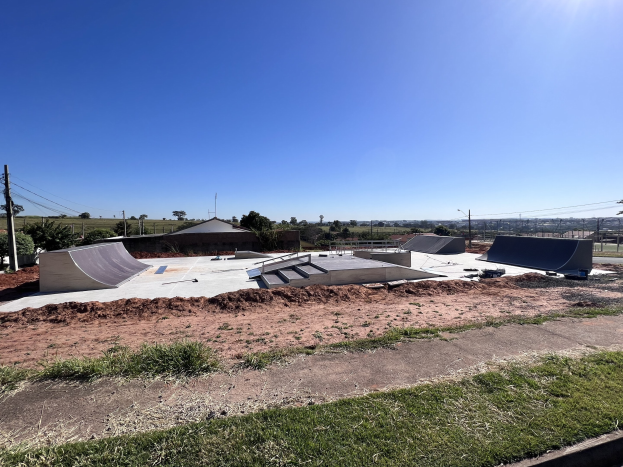 Skate-Anlage mit mehreren Rampen, Gras, Bäumen, Pfählen, Drähten und einem klaren blauen Himmel.