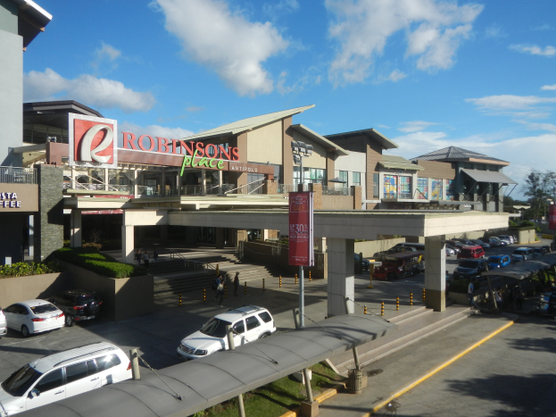 Ein belebter Einkaufszentrum mit verschiedenen Gebäuden, Geschäften, Fahrzeugen auf der Straße, Fußgängern auf dem Gehweg, Straßenlaternen, Informationsschildern, Grünflächen und einem bewölkten Himmel.