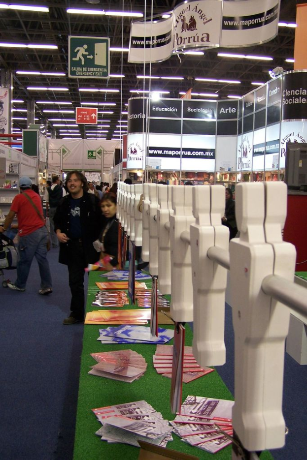 Menschen stehen und gehen in einem Raum mit Tischen, auf denen Ausrüstung, Bücher und Papiere liegen, und Ständen mit Text, Schildern und Bannern im Hintergrund, unter einer Decke.