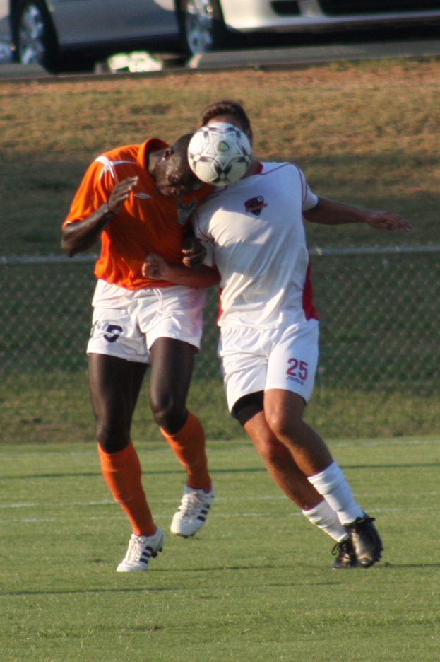 Zwei Spieler auf einem umz√§unten Platz, einer in einem orangen T-Shirt, der den Ball mit dem Kopf sch√§gt, mit Autos und Gras im Hintergrund.