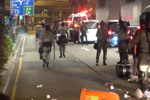 Eine Gruppe von Polizisten in Helmen, die Waffen halten, geht öffentlich mit Fahrzeugen, Pflanzen, einem Zaun und Schildern auf der linken Seite und beleuchteten Gebäuden im Hintergrund.