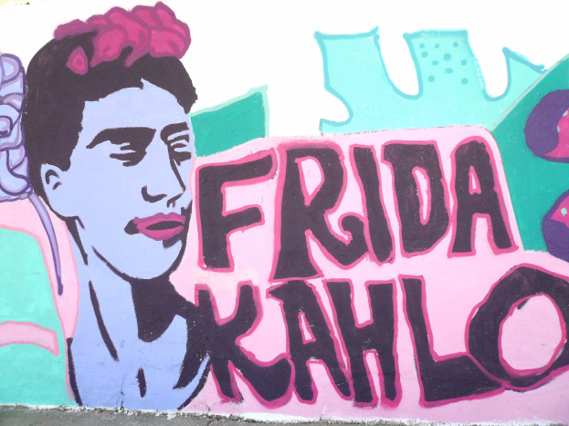 Ein leuchtendes Frida-Kahlo-Mosaik auf einem Gebäude in Mexiko-Stadt, das ihr Porträt mit einer Blume im Haar und den Text 'Fridakahlo' in fetter, schwarzer Schrift zeigt.