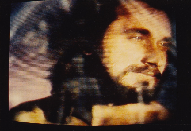 Mann mit Bart und Schnurrbart in einem weißen Hemd, der direkt in die Kamera schaut und einen ernsten Gesichtsausdruck hat, vor einem dunklen Hintergrund.