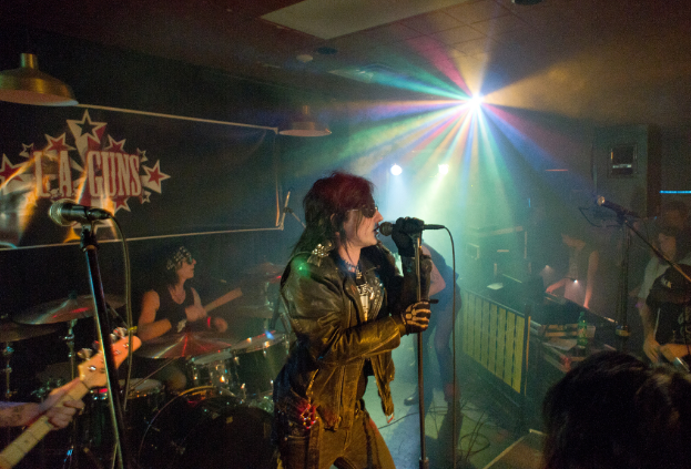 Eine Frau in einer schwarzen Jacke singt in ein Mikrofon auf der Bühne, mit einem Mann in Sonnenbrille und Stirnbinde, der ein Instrument spielt, im Hintergrund und einem "LA Guns"-Plakat und Diskoleuchten.
