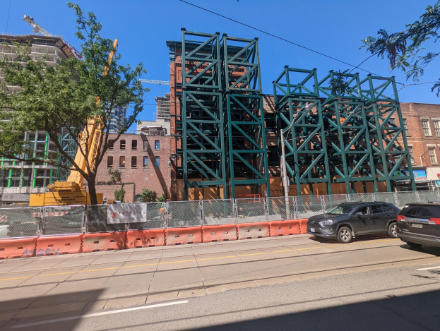 Baustelle mit teilweise errichteter Metallbrücke im Vordergrund, Autos am Straßenrand geparkt, ein Zaun, Bäume, Gebäude mit Fenstern, ein Kran und ein Himmel im Hintergrund.