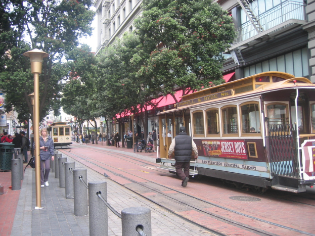 Eisenbahnschienen verlaufen entlang einer Stra{\ss}e mit Z"ugen, auf denen Text angezeigt wird, einer Person in der N"he, kleinen Masten mit Seilen, Stra{\ss}enlaternen, B"umen, vielen Menschen, einem M"llner auf der linken Seite und Geb"uden mit Leitern auf der rechten Seite.