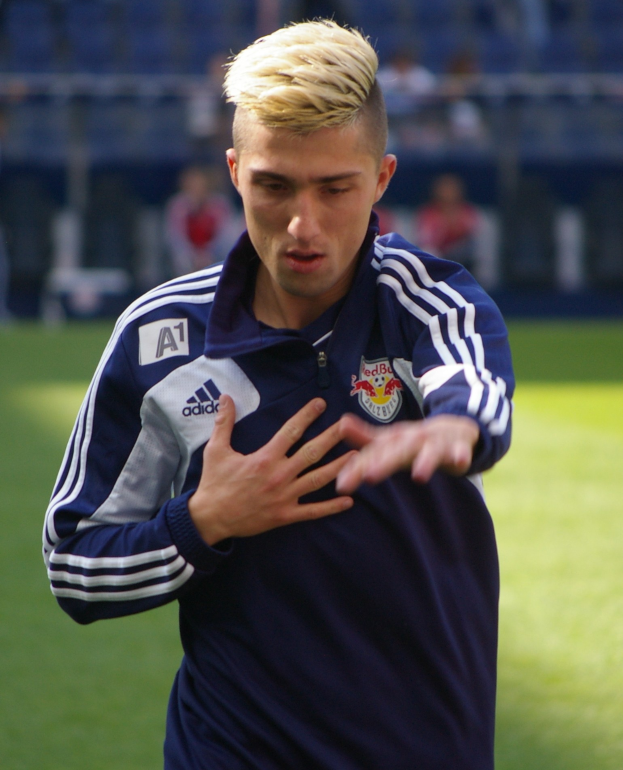 Ein junger Mann in einem blauen T-Shirt steht auf einem Fußballfeld und streckt die Arme aus, mit entschlossener Miene.