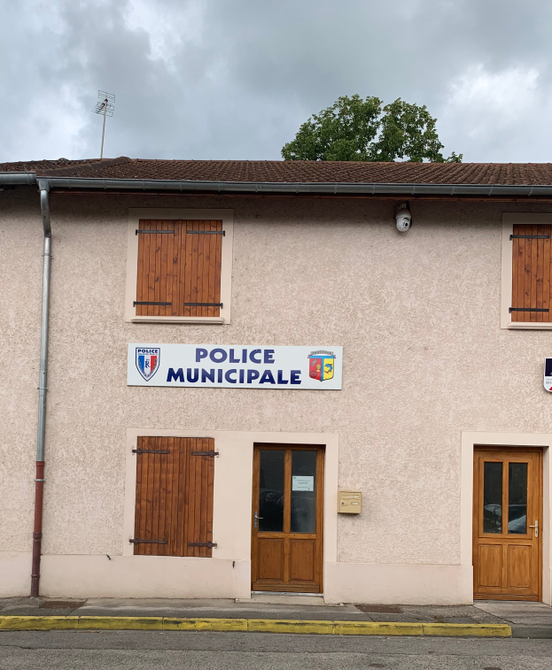 Ein Polizeigebäude mit einem Schild, Fenstern, Türen, einer Namensplakette, einem Rohr, einem Baum und einem bewölkten Himmel im Hintergrund.