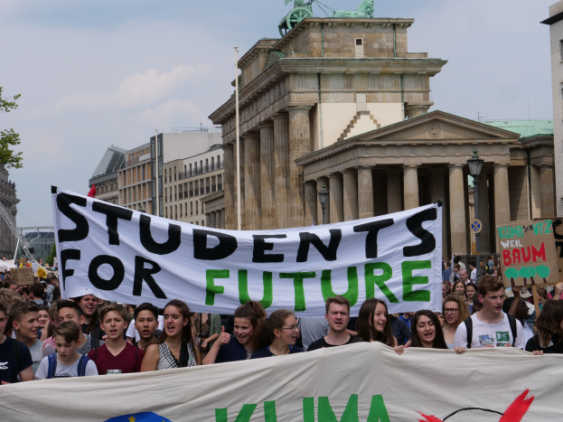 Gruppe von Schülern marschiert in Berlin mit einem leuchtend bunten "Students for Future"-Schild vor einem Hintergrund aus Gebäuden, Bäumen und Himmel.