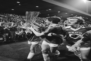 Eine Gruppe von Männern, die einen Pokal auf einem Fußballfeld halten, umgeben von einer Menge hinter einem Zaun, die ihren Sieg im FA-Cup-Finale feiern, dargestellt in Schwarz-Weiß.
