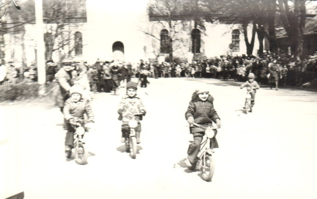 Schwarze und weiße Aufnahme von Kindern, die Fahrräder eine Straße hinunterfahren, mit einem Gebäude und Bäumen im Hintergrund.