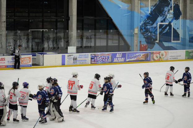Gruppe junger Menschen beim Eisschnellaufen auf einer Indoor-Eisbahn, die Helme tragen und Stöcke halten, mit einer Person links daneben und Bannern, Säulen und Glaswänden im Hintergrund.