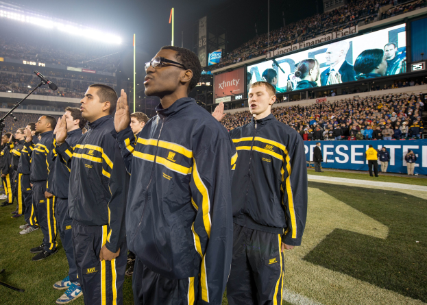 Eine Gruppe von Männern in blauen und gelben Jacken steht auf einem Fuballfeld, mit einer Menge, Schildern, Fahnen und Stadionbeleuchtung im Hintergrund, sowie Mikrofonen, die eine Marschkapellenaufführung andeuten.