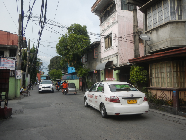 Ein weißes Taxi fährt auf einer Straße mit hohen Gebäuden entlang, mit Menschen auf Fahrrädern, anderen Fahrzeugen, Strommasten mit Drähten, Bäumen und einem klaren blauen Himmel im Hintergrund.