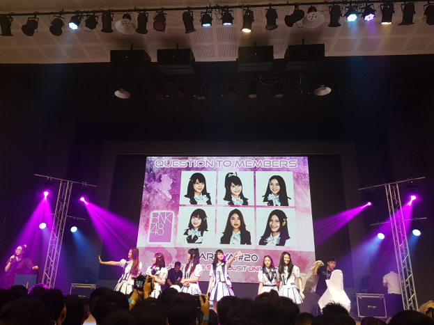 Eine Gruppe von Menschen, die auf einer hell erleuchteten Bühne performen, mit einer Menge darunter, mit einem Sänger mit Mikrofon und einem Musiker, der ein Instrument spielt, während ein großes Display Bilder der K-Pop-Gruppe BK48 zeigt.