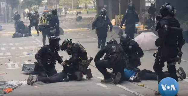 Eine Gruppe von Polizisten in Einsatzausrüstung steht um einen verletzten Mann auf dem Boden auf einer Straße, mit Bäumen und Gebäuden im Hintergrund.