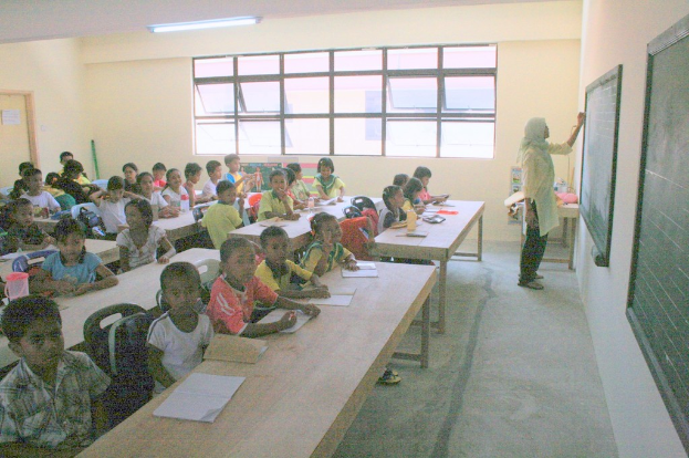 Ein Klassenzimmer mit einer Frau, die an einer Tafel rechts schreibt, Fenstern in der Mitte und einer Lampe an der Decke, mit Tischen voller Bücher und Wasserflaschen sowie Stühlen mit Kindern darauf.