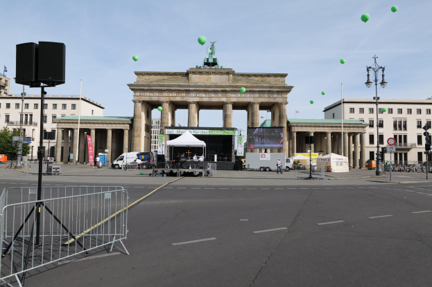 Das Brandenburgische Tor in Berlin, Deutschland, mit seinen ikonischen Säulen und Statuen, umgeben von einer festlichen Atmosphäre für den Berlin-Marathon, mit Gebäuden, Laternenmasten, Verkehrssignalen, Zelten, Fahrzeugen, Menschen, Fahrrädern und grünen Luftballons im bewölkten Himmel.