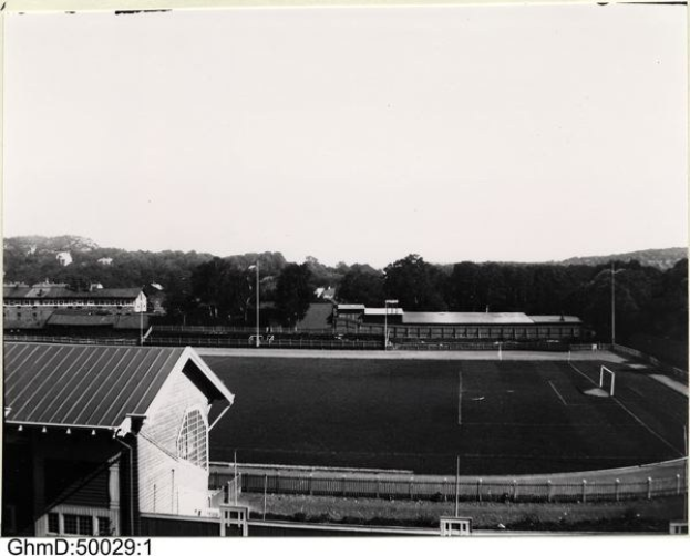 Schwarzes und weißes Foto eines Fußballfeldes mit einem Schuppen auf der linken Seite, einem umgebenden Zaun, zentralen Pfosten und Bäumen mit Himmel im Hintergrund, beschriftet mit "Gillingham Football Club, 1960er Jahre" unten.