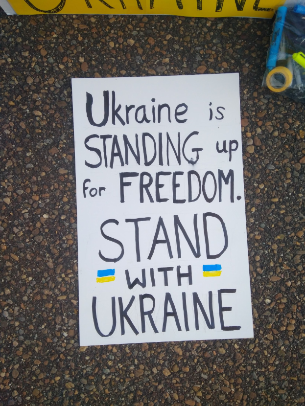 Weißes Papier mit "Ukraine steht für Freiheit, steht mit Ukraine" Text auf dem Boden vor einem gelben Banner, mit einer Plastikabdeckung mit verschiedenen Gegenständen rechts.