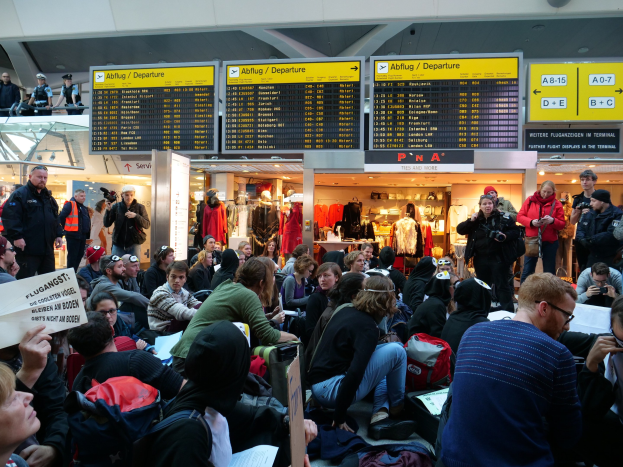 Eine große Gruppe von Menschen sitzt und steht in einem Flughafen während einer Protestaktion, wobei einige Taschen und Papiere halten, während im Hintergrund Schilder mit Text, Schaufensterpuppen in Kleidern und Deckenleuchten zu sehen sind.