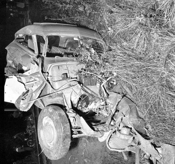 Schwarz-weiß-Foto eines schwer beschädigten Autos mit eingedellter Front und zersplitterten Fenstern, das auf der Seite der Straße mit einem Baum und verstreuten Gegenständen im Hintergrund liegt.