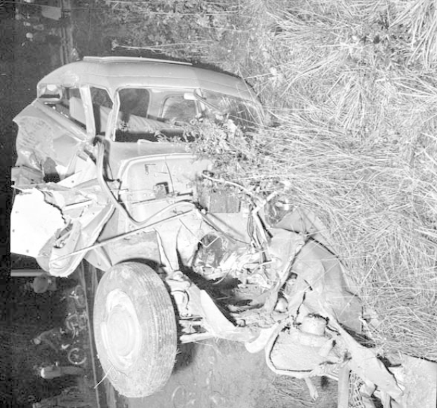 Schwarz-weiß-Foto eines schwer beschädigten Autos mit eingedellter Front und zersplitterten Fenstern, das auf der Seite der Straße mit einem Baum und verstreuten Gegenständen im Hintergrund liegt.