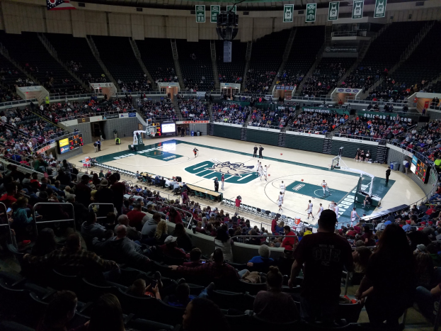 Eine große Halle mit Zuschauern auf den Tribünen, die ein Basketballspiel auf einem zentralen Platz beobachten, mit Anzeigetafeln und Beleuchtung oben.