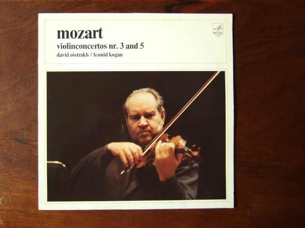 Ein Mann in einem schwarzen Mantel spielt Geige auf einem Holztisch, mit einem schwarzen Hintergrund und einer kleinen Inschrift oben auf dem Bild.