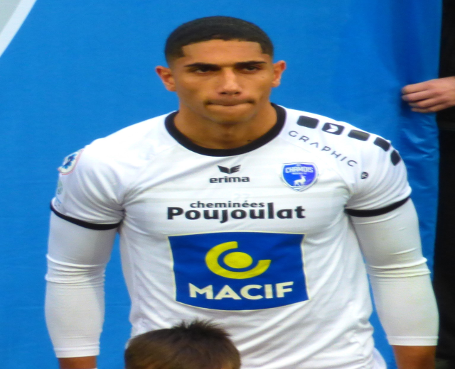 Ein Fußballspieler in einem weißen T-Shirt steht auf einem Feld mit verschränkten Armen und entschlossener Miene neben einem anderen Mann mit einem blauen Tuch im Hintergrund.