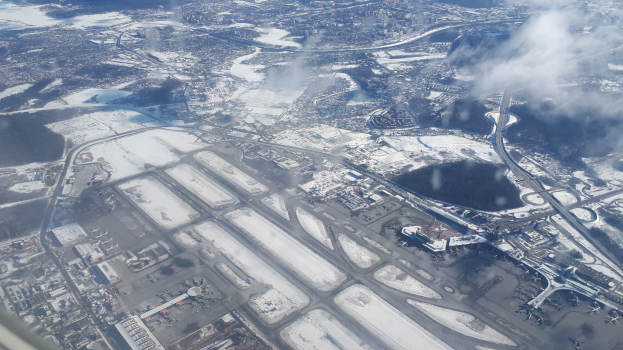 Eine Luftaufnahme eines Flughafens mit schneebedecktem Boden, Gebäuden, Straßen, Brücken, Wasser, Bäumen und Wolken am Himmel.