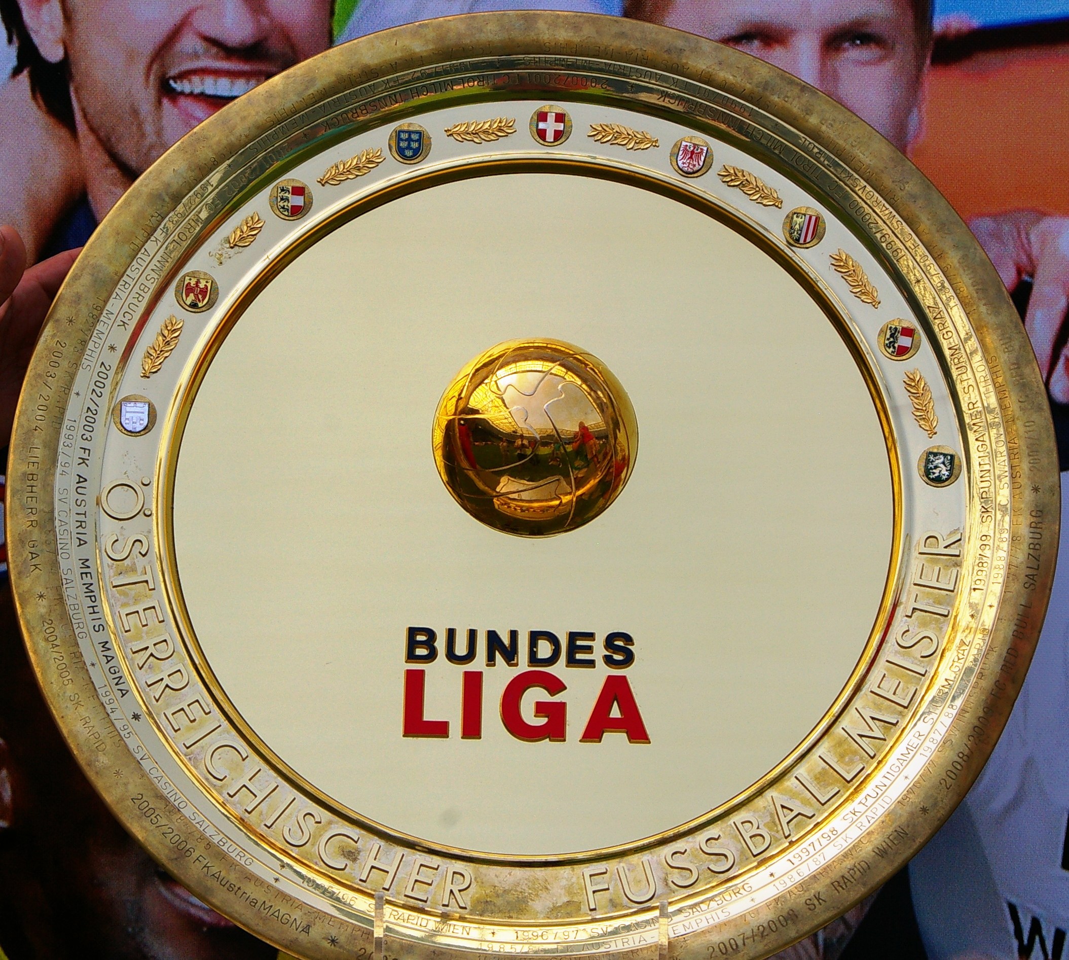 Mann hält goldenen Schild mit 'Bundesliga'-Text, im Hintergrund ein Banner mit drei Personen
