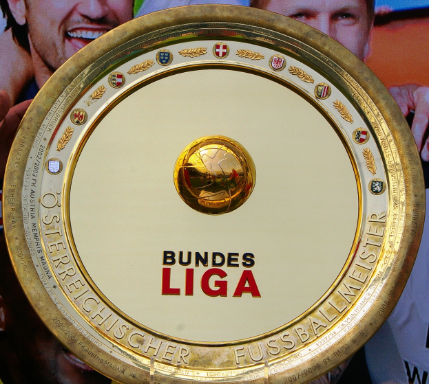 Mann hält goldenen Schild mit 'Bundesliga'-Text, im Hintergrund ein Banner mit drei Personen
