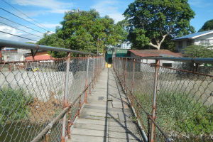 Eine Hängebrücke spannt einen Fluss, flankiert von Zäunen, mit üppiger Vegetation, fernen Häusern und einem klaren blauen Himmel im Hintergrund.