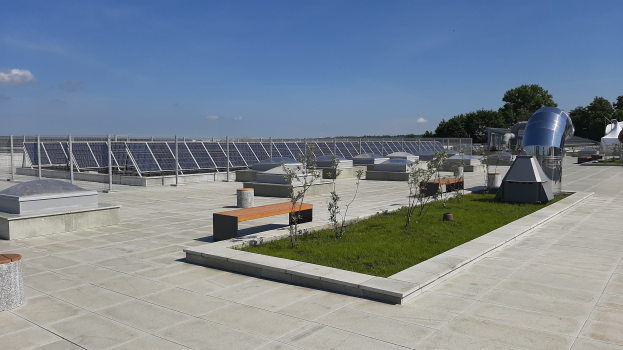 Ein Gebäude mit Solar-Panelen auf dem Dach, umgeben von Bänken, Pflanzen, Gras und Bäumen unter einem bewölkten Himmel.
