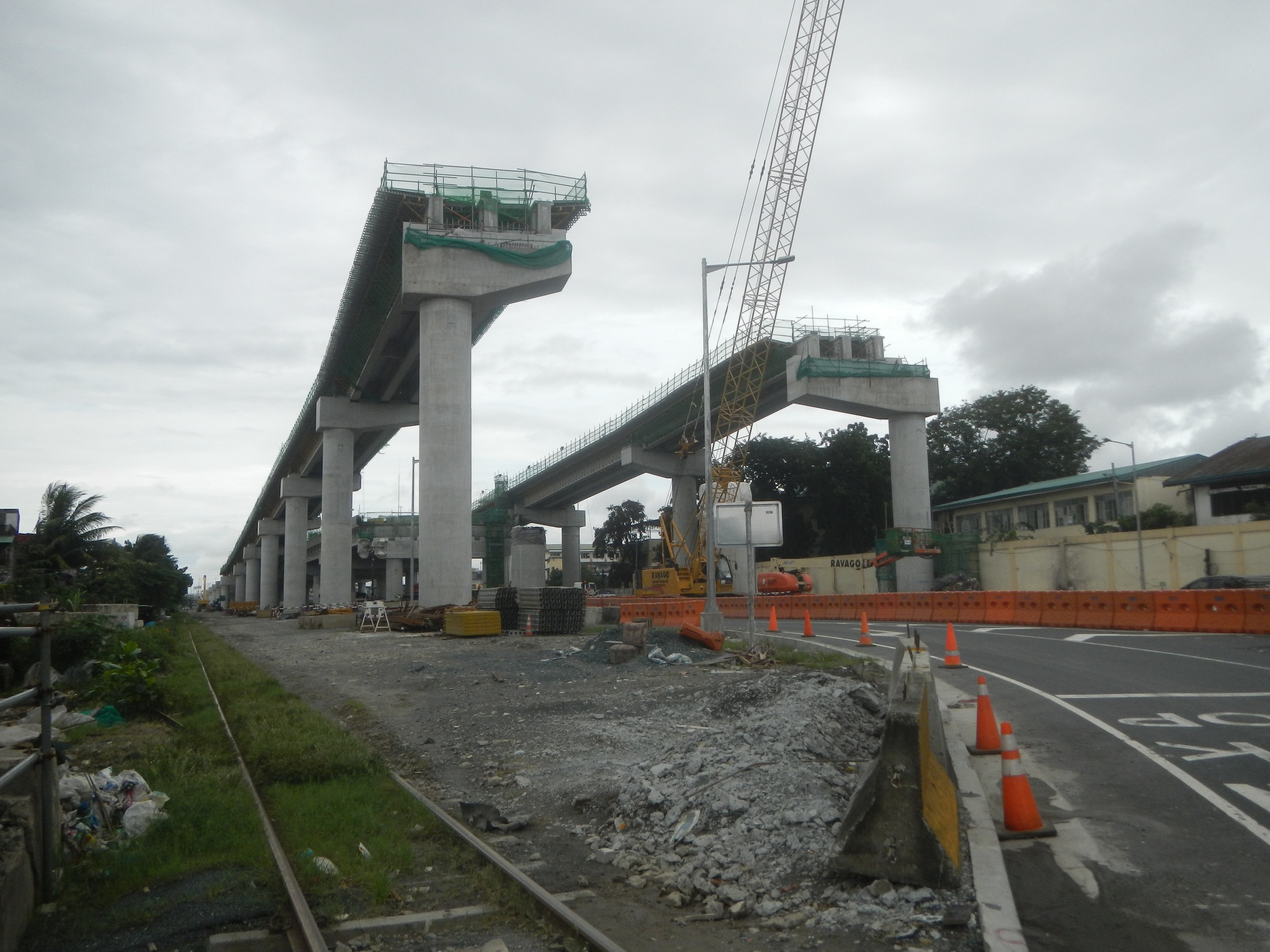 Baustelle mit einer Brücke im Hintergrund, Straße mit Verkehrskegeln markiert, verstreute Steine und Gras, Eisenbahnschiene links, Bäume und Gebäude flankieren die Straße und ein bewölkter Himmel.