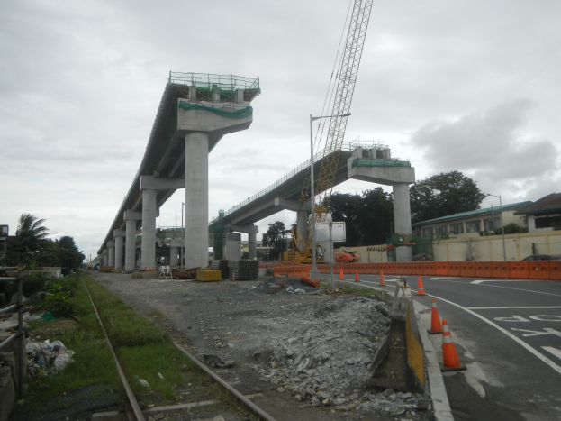 Baustelle mit einer Brücke im Hintergrund, Straße mit Verkehrskegeln markiert, verstreute Steine und Gras, Eisenbahnschiene links, Bäume und Gebäude flankieren die Straße und ein bewölkter Himmel.