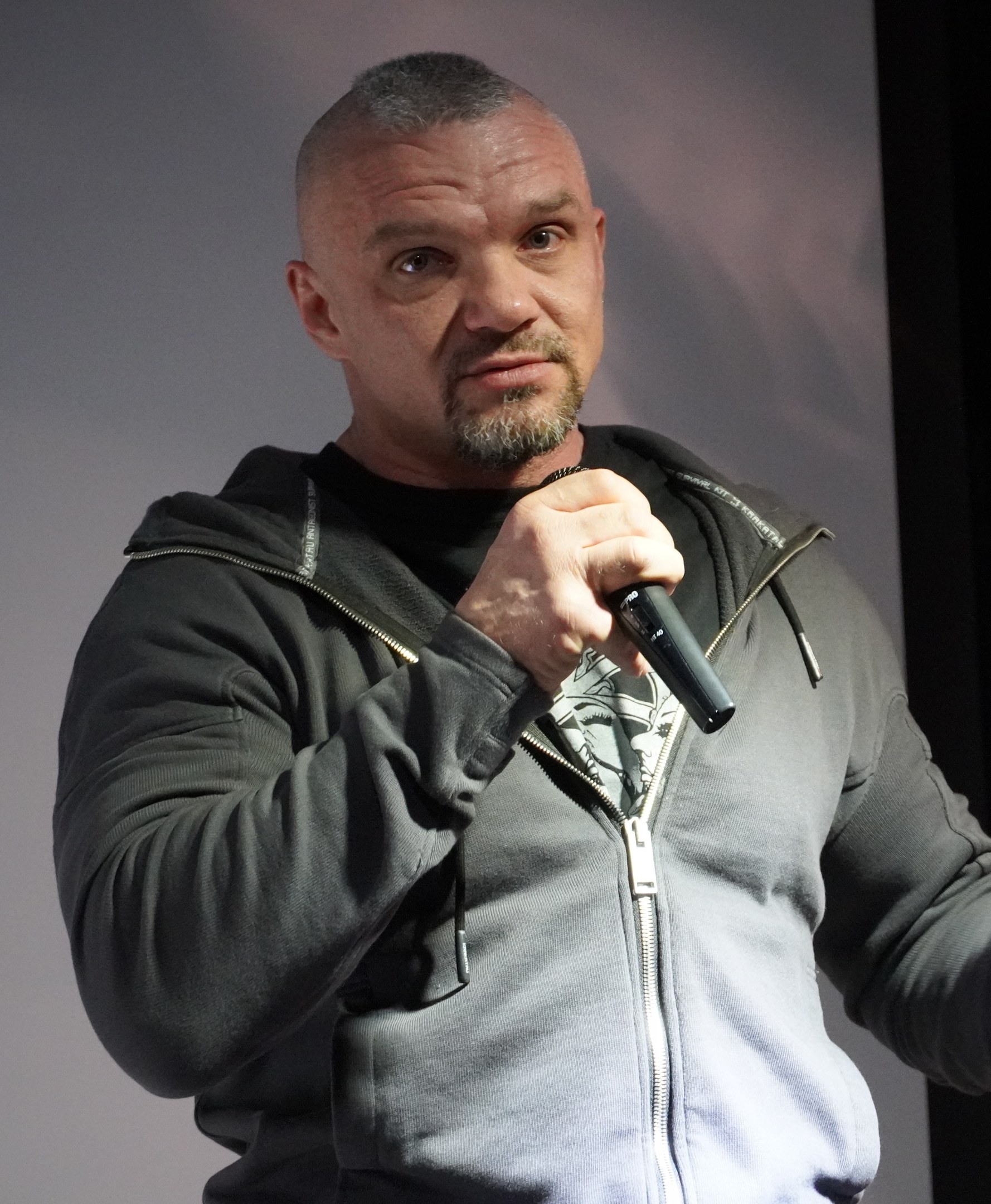 Ein Mann in einer schwarzen Kapuzenjacke und einem schwarzen Anzug mit einem wei├čem Hemd, der ein Mikrofon h├Ąlt und selbstbewusst vor einer Wand steht.