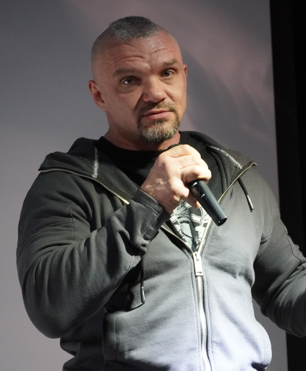 Ein Mann in einer schwarzen Kapuzenjacke und einem schwarzen Anzug mit einem wei├čem Hemd, der ein Mikrofon h├Ąlt und selbstbewusst vor einer Wand steht.