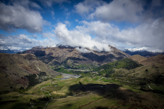 Eine Totalenansicht einer bergigen Landschaft in Queenstown, Neuseeland, mit saftig grünem Gras, verstreuten Bäumen, einer gewundenen Straße und einem Himmel voller weißer, flauschiger Wolken.