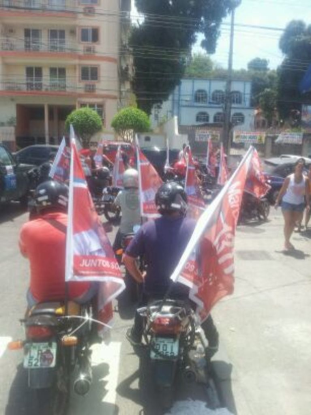 Eine Gruppe von Menschen, die Fahrräder fahren und eine Fahne halten, mit Gebäuden, Autos, Bäumen, Bannern und einem bewölkten Himmel im Hintergrund.