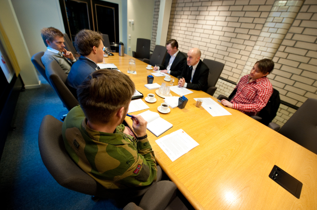 Eine Gruppe von Menschen, die um einen Tisch mit Tassen, Büchern, Papieren, einer Flasche, einem Krug und Stiften während einer Besprechung sitzen.