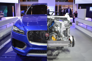 Ein blauer Jaguar F-Pace R-Dynamic S AWD wird auf der IAA in Frankfurt präsentiert, umgeben von Menschen, die das Auto beobachten und fotografieren, und Bildschirme im Hintergrund.