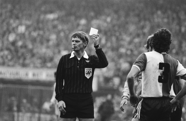 Schwarzes und weißes Bild eines Schiedsrichters mit roter Karte vor einer Menge von Fußballspielern, mit einem Zaun im Hintergrund.