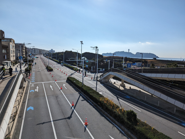 Eine Straße mit Verkehrskegeln, eine Brücke mit Geländern, Laternenpfähle, Gebäude mit Fenstern und einige Menschen, vor einem Hügel- und Wolkenhimmel als Teil des Takayama-Brücken-Projekts.